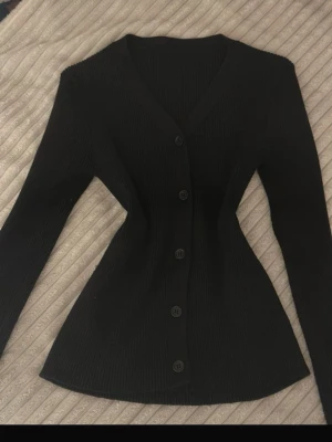 Svart ribbad kofta med knappar - Snygg svart ribbad kofta med v-ringning och långa ärmar. Koftan har en figurnära passform och stängs med fem svarta knappar framtill. Perfekt att styla med jeans eller kjol för en clean look.