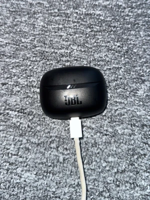 JBL Tune Beam 2 Trådlösa Hörlurar - JBL Tune Beam 2 trådlösa hörlurar med laddningsetui. Svart färg, USB-C laddning. Hörlurarna och etuiet ser ut att vara i gott skick med minimalt slitage. Perfekt för musik, samtal och träning med bekväm passform och bra ljudkvalitet.