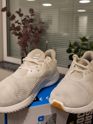 Vita Adidas sneakers med snörning 42 2/3 - Säljer ett par fräscha vita Adidas sneakers med mesh-ovandel och snygga detaljer i syntet. Skorna har rund tå, platt sula och klassisk snörning. Adidas-loggan syns på både plös och häl. Perfekta för dig som gillar stilrena och sportiga skor.