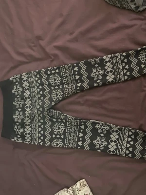 Svart loungewear-set med snöflingor - Mysigt loungewear-set i svart med vita snöflingor och klassiskt vintermönster. Setet består av långärmad tröja och byxor med resår i midjan. Perfekt för kalla dagar hemma. Materialet känns mjukt och stretchigt, troligen bomulls- eller polyesterblandning.