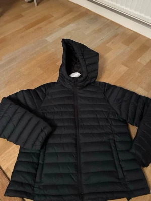 Svart pufferjacka med huva motsvarar stl M  - Säljer en svart pufferjacka med huva och dragkedja framtill. Jackan har två sidofickor och quiltad design med horisontella sömmar. Perfekt för kalla dagar och riktigt skön att bära. Materialet är syntet och jackan har en klassisk, enkel look. Står L men tycker den är liten o storleken :)