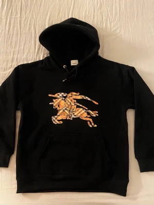 Svart hoodie med Burberry-ryttare - Svart hoodie från Burberry med stor broderad ryttare på häst i klassiskt beige-rutigt mönster på bröstet. Hoodien har huva med dragsko och en stor magficka. Tillverkad i mjukt bomullsmaterial och har ribbade muddar vid ärmslut och nederkant.