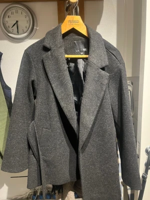 Mörkgrå rock från 157, storlek XS - Stilren mörkgrå rock från 157 med klassisk krage och snyggt fall. Rocken har två sidofickor och är helfodrad med svart innerfoder. Perfekt för dig som gillar en enkel och tidlös look.