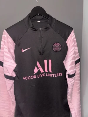 PSG träningströja svart/rosa Nike - Snygg långärmad träningströja från PSG i svart med rosa detaljer och loggor. Tröjan har halv dragkedja, rosa mönstrade ärmar och klubbmärke på bröstet. Materialet är troligtvis polyester, perfekt för sport och träning.
