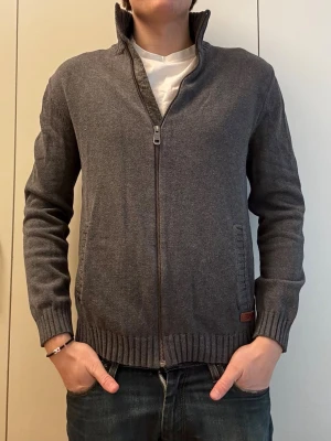 Mörkgrå ziptröja från Esprit - Mörkgrå stickad ziptröja från Esprit med hög krage och ribbade muddar. Tröjan har två fickor framtill och en diskret Esprit-logga vid nederkanten. Perfekt för lager på lager och enkel att matcha med olika outfits. Längd: 62cm, P2P: 42cm