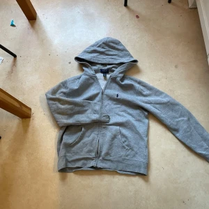 Grå zip hoodie från Polo Ralph Lauren - Klassisk grå hoodie med dragkedja från Polo Ralph Lauren. Hoodien har huva, två fickor framtill och den ikoniska lilla broderade loggan på bröstet. Perfekt för en chill och avslappnad stil. Tillverkad i mjukt bomullsmaterial.