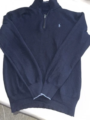 Mörkblå halvzip från Ralph Lauren - Mörkblå stickad tröja från Ralph Lauren med halv dragkedja och klassisk broderad logga på bröstet. Tröjan har hög krage och ribbade muddar vid ärmslut och nederkant. Perfekt för en stilren och avslappnad look.
