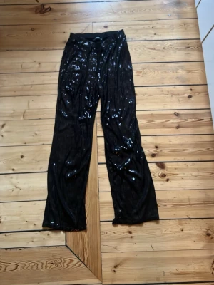 Svarta paljettbyxor med vida ben - Snygga svarta byxor med paljetter som ger ett glittrigt intryck. Byxorna har vida ben och resår i midjan för skön passform. Perfekta för dig som vill sticka ut och addera lite extra bling till din outfit.