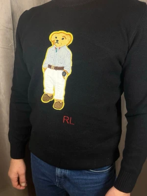 Svart stickad tröja med björn från Ralph Lauren - Svart stickad tröja från Ralph Lauren med ikonisk broderad björn på bröstet. Björnen har skjorta, vita byxor och bruna skor. RL-broderi i rött under motivet. Klassisk rund hals och ribbade muddar.