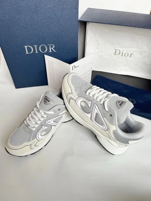 Gråa Dior sneakers med vit detalj - Snygga sneakers från Dior i grått mesh och mocka med vita och ljusgrå detaljer. Klassisk låg modell med tjock sula och vita skosnören. Stora Dior-loggor på sidan och tungan ger en exklusiv vibe. Perfekta för dig som vill sticka ut med stilrena och trendiga skor.
