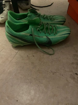 Gröna Nike Mercurial fotbollsskor - Säljer ett par grymma gröna Nike Mercurial fotbollsskor med svarta detaljer och snörning. Skorna har en sportig design med tunna ränder och är perfekta för dig som vill sticka ut på planen. Lätta och smidiga för snabba rörelser.