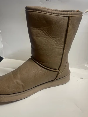 Bruna höga tofflor i skinnimitation - Säljer ett par bruna, höga tofflor i skinnimitation med mjuk insida. De har en enkel design utan synliga märken och en rund tå. Perfekta för att hålla fötterna varma hemma under kyliga dagar.