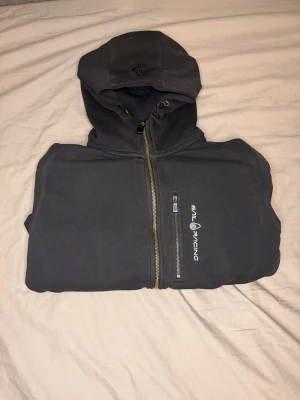 Svart hoodie från Sail Racing - Svart hoodie med dragkedja från Sail Racing. Har huva med dragsko, två sidofickor och en bröstficka med dragkedja och logga. Perfekt för en avslappnad och sportig stil.