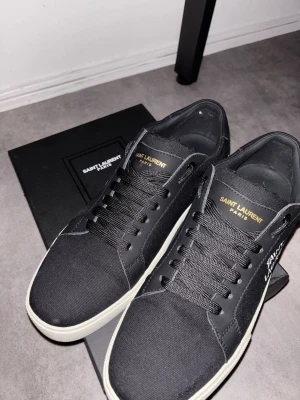 Svarta Saint Laurent sneakers - Snygga svarta sneakers från Saint Laurent med vit sula och broderad logga på sidan. Skorna har svarta skosnören, diskret guldfärgad text på plösen och är tillverkade i canvas och läderdetaljer. Perfekta för dig som gillar stilrena och exklusiva sneakers.