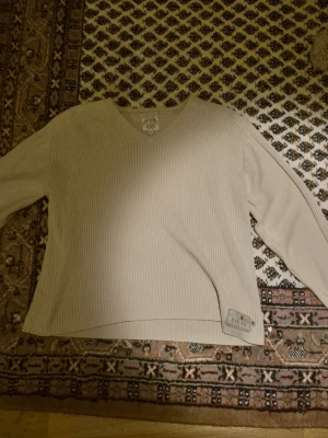 Beige ribbad tröja med V-ringning - Snygg beige ribbad tröja med V-ringning och långa ärmar. Tröjan har en avslappnad passform och en liten patchdetalj nertill. Perfekt för dig som gillar stilrena och enkla plagg. Köptes secondhand, inte använts sedan köp.