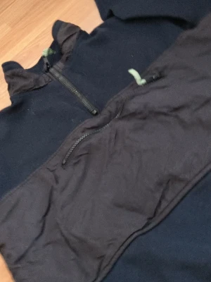 Svart hoodie med dragkedja och ficka - Svart hoodie med halv dragkedja framtill och stor magficka med dragkedja. Huva med snörning i kontrastfärg. Perfekt för en avslappnad streetwear-look. Materialet ser ut att vara en mix av bomull och polyester.