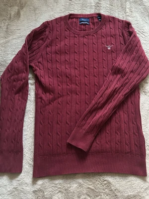 Röd kabelstickad tröja från GANT - Säljer en röd kabelstickad tröja från GANT med broderad logga på bröstet. Tröjan har rund halsringning, ribbade muddar och är tillverkad i mjuk bomull. Perfekt för dig som gillar klassisk och stilren look. Den är storlek M men den är stretch och den sitter perfekt på en som är 170! Pris kan diskuteras vid snabb affär!