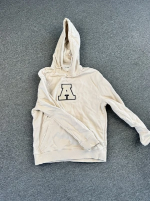 Beige hoodie från Axel Arigato - Snygg beige hoodie från Axel Arigato med stor svartvit A-logga på bröstet. Hoodien har huva med dragsko och en klassisk känguruficka framtill. Tillverkad i mjukt bomullsmaterial som är skönt att ha på sig. Perfekt för en avslappnad och trendig look.