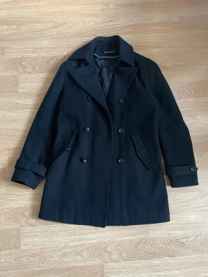 Svart rock från Bondelid - Klassisk svart rock från Bondelid med dubbelknäppning, bred krage och två snedställda fickor. Kavajen har quiltat innerfoder och dekorativa knappar vid ärmsluten. Storlek s. Kom gärna med frågor:)