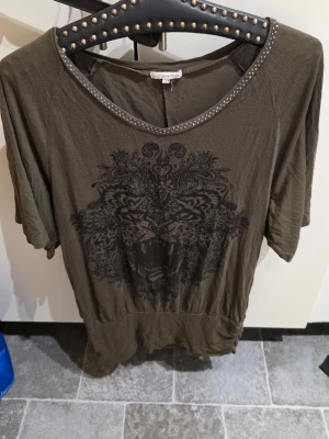 Olivgrön topp med tigertryck - Cool olivgrön topp med stort svart tigertryck framtill och nitar längs den runda halsringningen. Modellen har korta ärmar och avslappnad passform, perfekt för en edgy look.