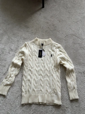 Vit kabelstickad tröja Polo Ralph Lauren storlek M passar S - Kabelstickad vit tröja från Polo Ralph Lauren med halv dragkedja och ribbad krage. Tröjan har långa ärmar, ribbade muddar och den klassiska broderade loggan på bröstet. Perfekt för en clean och stilren look.