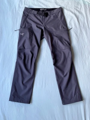 Mörkgrå cargobyxor från Arc'teryx - Snygga mörkgrå cargobyxor från Arc'teryx med raka ben och flera praktiska fickor med dragkedja. Tillverkade i ett slitstarkt, stretchigt material som passar perfekt för äventyr och vardag. Broderad logga på låret ger en clean outdoor-look.