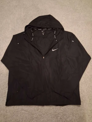 Svart Nike vindjacka med huva - Snygg svart vindjacka från Nike med dragkedja och huva. Jackan har Nike-logga på bröstet och texten 'Nike Running' på insidan av huvan. Perfekt för löpning eller när du vill ha en lätt jacka. Tillverkad i tunt, vindtätt material.