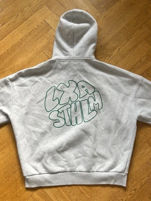 Grå hoodie från LXA STHLM - Snygg grå hoodie från LXA STHLM med stor grön logga i bubbelstil på ryggen. Klassisk passform med huva och dragsnören samt ribbade muddar vid ärmar och midja. Tillverkad i mjukt sweatshirtmaterial, perfekt för en chill och avslappnad stil.