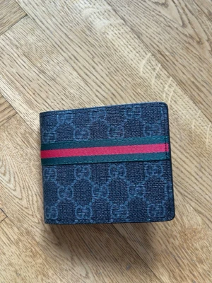 Gucci plånbok med GG-mönster - Snygg svart plånbok från Gucci med klassiskt GG-mönster och en ikonisk röd-grön rand över framsidan. Plånboken är i canvasmaterial med detaljer i läder och har en stilren, fyrkantig form. Perfekt för dig som gillar exklusiva accessoarer.