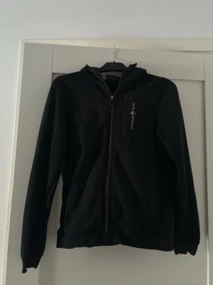 Svart hoodie från Sail Racing - Svart hoodie från Sail Racing med dragkedja framtill och en bröstficka med dragkedja. Märkets logga är tryckt på bröstet. Hoodien har huva och är tillverkad i ett mjukt material som känns skönt mot huden. Perfekt för en avslappnad och sportig stil.