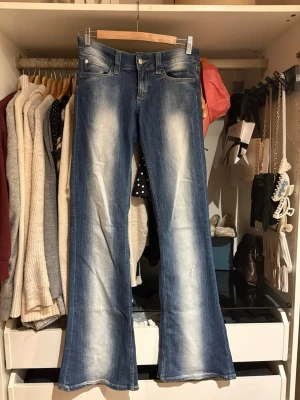 Blå bootcut jeans med slitningar - Säljer ett par blå bootcut jeans med snygga slitningar och ljusa partier framtill. Jeansen har klassisk femficksmodell, normal midja och dragkedja med knapp. Perfekta för dig som gillar retrovibbar och vill ha ett par jeans som sticker ut.