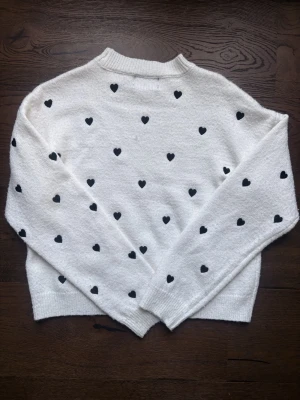 Vit tröja med svarta hjärtan - Superfin vit tröja med svarta hjärtan över hela. Tröjan har en rak passform, ribbad halsringning och muddar vid ärmslut och nederkant. Perfekt för dig som gillar gulliga detaljer och en mysig stil.