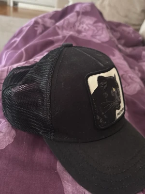 Svart truckerkeps med pantermärke - Snygg svart truckerkeps från The Farm med mesh på sidorna och ett broderat märke med en svart panter framtill. Kepsen har böjd skärm och justerbar baksida. Perfekt för en cool och avslappnad stil.