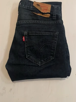 Mörkblå Levi's jeans med klassisk ficka - Säljer ett par mörkblå jeans från Levi's med den ikoniska röda loggan på bakfickan och klassisk femficksdesign. Jeansen har raka ben och är tillverkade i slitstark denim. Perfekta för dig som gillar tidlös och enkel stil.
