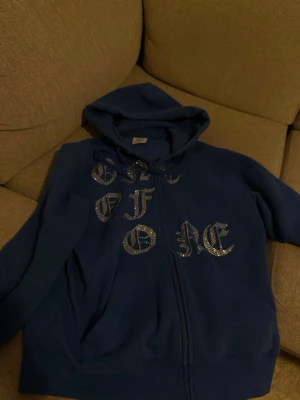 Blå hoodie från One of One - Säljer en blå hoodie från One of One med dragkedja och huva. Framsidan har stora bokstäver i glittrande stenar som ger en unik och trendig look. Hoodien har fickor framtill och är gjord i mjukt material. Original pris 1.500kr