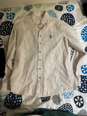Vit mönstrad skjorta från U.S. Polo Assn. - Säljer en vit skjorta från U.S. Polo Assn. med brunt abstrakt mönster och klassisk krage. Skjortan har knappar hela vägen och en liten broderad logga på bröstet. Perfekt för dig som gillar stilrena detaljer och skön passform.
