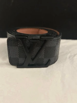 Louis Vuitton rutigt svart bälte - Snyggt svart bälte från Louis Vuitton med klassiskt rutigt mönster och stor LV-logga i metall som spänne. Bältet har en exklusiv känsla och är gjort i skinn med en mörk finish. Perfekt accessoar för att lyfta din outfit.