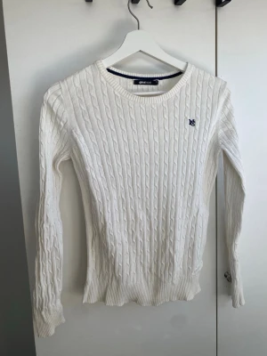 Vit kabelstickad tröja Gina Tricot - Kabelstickad vit tröja från Gina Tricot med rund halsringning och broderad blå logga på bröstet. Tröjan har långa ärmar och ribbade muddar vid ärmslut och nederkant. Perfekt för en stilren och klassisk look.