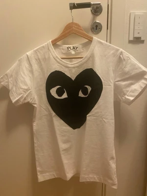 Vit t-shirt Comme des Garçons Play  - Snygg vit t-shirt från Comme des Garçons Play i storlek S. Framsidan har ett stort svart hjärta med ögon, vilket ger en unik och cool vibe. Klassisk rund hals och korta ärmar. Tillverkad i mjuk bomull för skön känsla hela dagen. Pris ej hugget i sten!