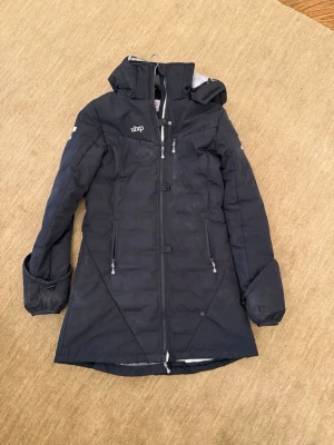 Svart lång parkas från Uhip - Snygg svart lång parkas från Uhip med huva och dragkedja framtill. Jackan har två stora fickor med dragkedja och justerbar midja. Perfekt för kalla dagar och har en stilren design med diskreta loggor på bröst och ärm. Uhip Ice Junior Long jacket. 