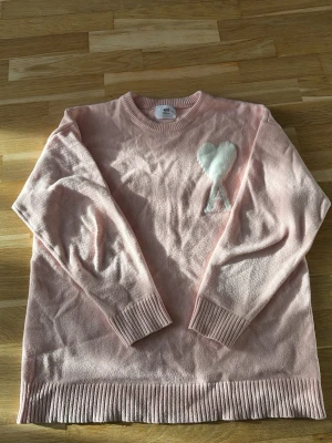 Ljusrosa stickad tröja med hjärta - Söt ljusrosa stickad tröja från AMI med ett vitt hjärta och bokstaven A broderat på bröstet. Tröjan har rund halsringning, långa ärmar och ribbade muddar vid ärmslut och nederkant. Perfekt för en mysig och trendig look.