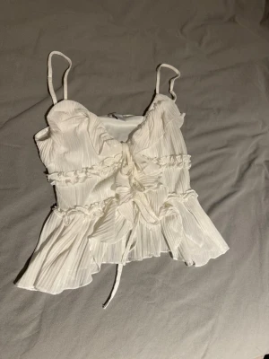 Vit plisserad linnetopp med volanger - Superfin vit linnetopp med tunna axelband, plisserat tyg och volangdetaljer. Toppen har snörning framtill och en luftig, flowy passform. 