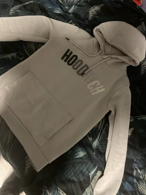 Beige hoodie från Hooch - Säljer en beige hoodie från Hooch med stor magficka och huva med dragsko. Framsidan har trycket 'HOODRICH' i svart och vitt. Hoodien har ribbade muddar vid ärmslut och nederkant. Perfekt för chill dagar och streetwear-stil.
