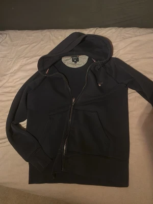 Gant kofta Mörkblå - Snygg mörkblå hoodie från GANT med dragkedja framtill och klassisk huva. Hoodien har två fickor och GANT-logga med broderad sköld på bröstet. Tillverkad i mjukt bomullsmaterial som känns skönt mot huden. Perfekt för en avslappnad och stilren look.