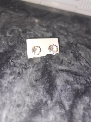 Silvriga örhängen med klar sten - Ett par små, silvriga örhängen med rund, klar sten i mitten. Klassisk och enkel design som passar till många stilar. Stenarna är infattade i metall och ger ett subtilt glittrande intryck.