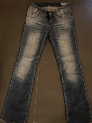 Blå bootcut jeans Lee - Snygga blå bootcut jeans från Lee med slitningar och ljus tvätt på låren och bakfickor. Klassisk femficksmodell med låg midja och Lee-logga på bakfickan. Perfekta för en avslappnad och trendig look. Waist 25 och längd 33!