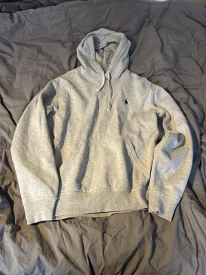 Grå hoodie från Polo Ralph Lauren - Snygg grå hoodie från Polo Ralph Lauren med klassisk huva och dragsko. Broderad logga på bröstet och stor känguruficka framtill. Mjuk insida och ribbade muddar vid ärmslut och nederkant. Perfekt för en avslappnad och stilren look.