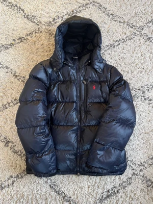 Svart pufferjacka från Ralph Lauren - Snygg svart/mörkblå pufferjacka från Ralph Lauren med huva och dragkedja framtill. Jackan har en röd broderad logga på bröstet och stora, fluffiga kanaler som ger en cool look. Perfekt för kalla dagar när du vill hålla stilen. Jackan är storlek xl. Mycket fint skick! 