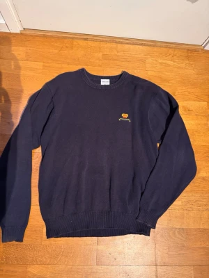 Mörkblå stickad tröja Bermuda - Mörkblå stickad tröja från Trimingham's Bermuda i 100% bomull. Tröjan har rund halsringning, ribbade muddar och en liten broderad krona med texten 'BERMUDA' på bröstet. Perfekt för dig som gillar klassisk och stilren design.