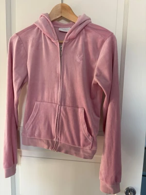 Rosa velourhoodie från Juicy Couture - Säljer en klassisk rosa velourhoodie från Juicy Couture med dragkedja framtill och broderad JC-logga på bröstet. Hoodien har huva, kängurufickor och ribbade muddar. Perfekt för en chill och trendig look.